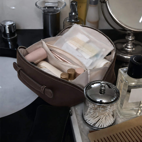 A dark brown zippered toiletry bag filled with cosmetics and accessories, placed on a bathroom counter beside a mirror, cotton swab jar, and perfume bottle.