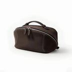 A dark brown makeup bag with a top handle, curved zipper, and white contrast stitching, shown on a clean white background.