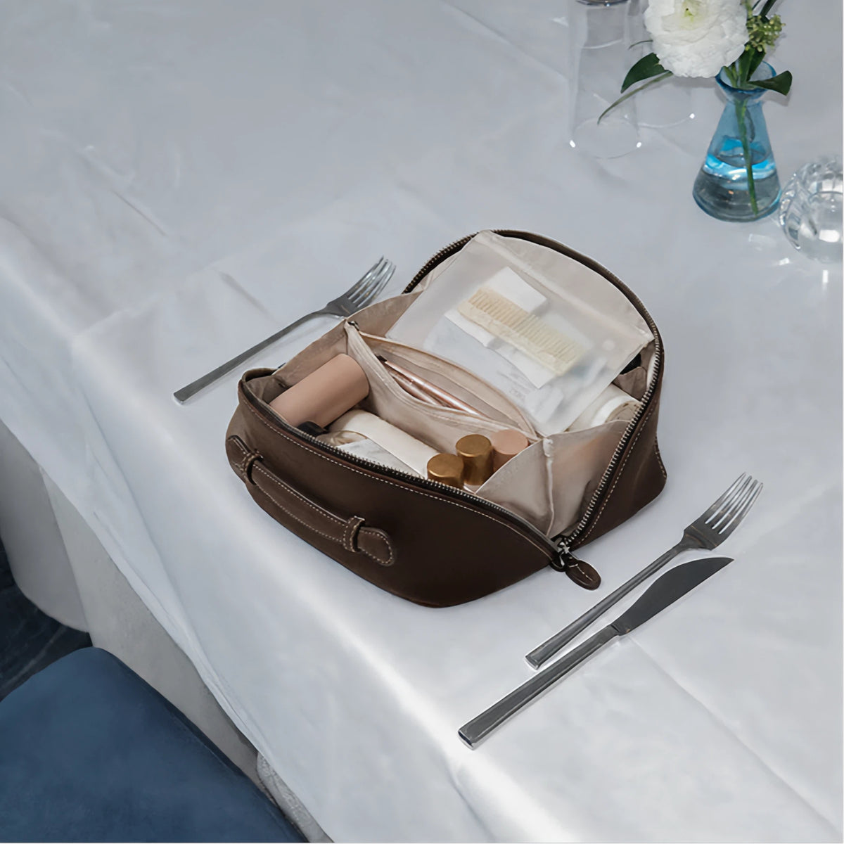 A dark brown zippered cosmetic pouch with a handle, shown open on a white tablecloth with cutlery and a vase of flowers in the background.