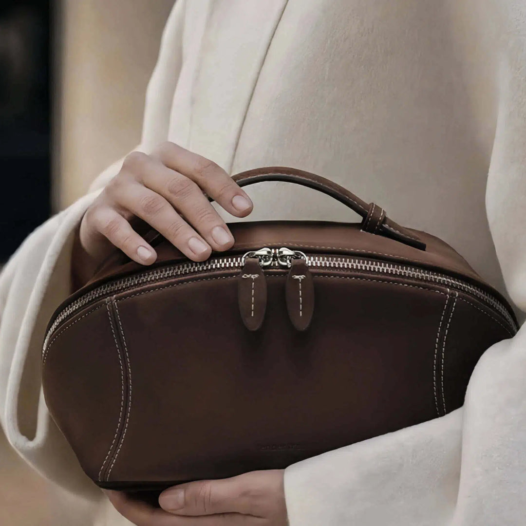 A person in a cream-colored coat holds a dark brown cosmetic bag with a top handle and silver-tone hardware.