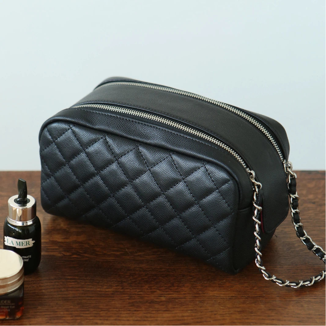 Black leather makeup bag with a chain detail on a wooden surface, next to a bottle and jar.