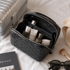 A black quilted cosmetic bag with a zippered open top, styled on a white fabric surface with travel props including a silver suitcase, skincare bottles, sunglasses, and a makeup brush.