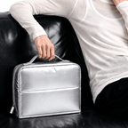 A person in a white long-sleeve top sits on a black leather couch and holds a shiny silver zippered cosmetic bag with a top handle.