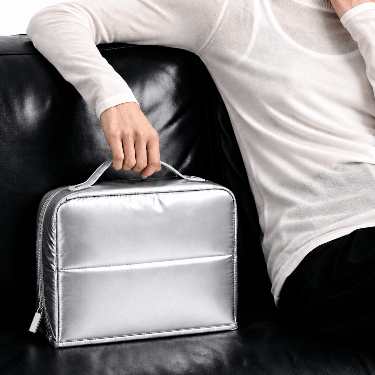 A person in a white long-sleeve top sits on a black leather couch and holds a shiny silver zippered cosmetic bag with a top handle.