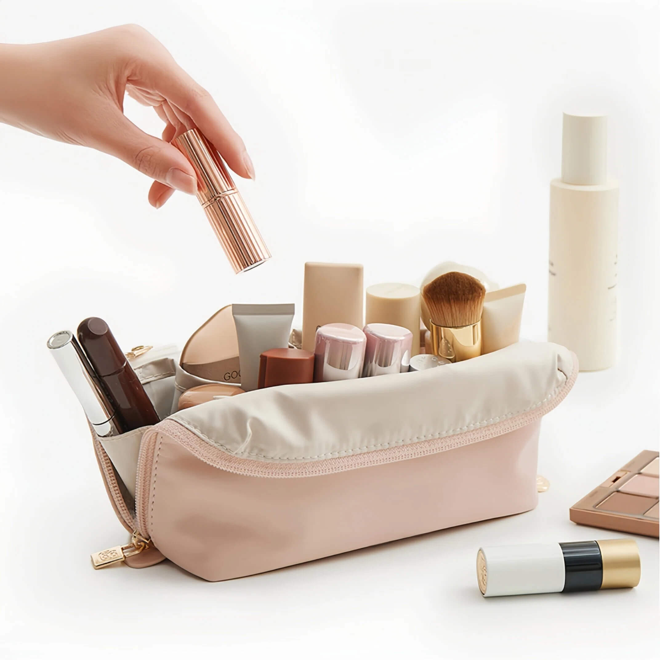 A pink makeup bag filled with cosmetics, shown on a clean white background with a hand placing a lipstick into the bag.