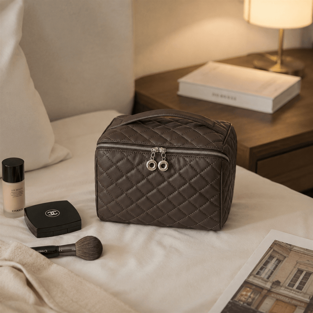 A quilted brown cosmetic bag with silver zipper pulls sits on a bed alongside makeup items and a brush, with a bedside lamp and book in the background.