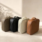 Four zippered toiletry or travel pouches in olive grey, black, cream, and brown, arranged in a row on a warm beige surface with soft shadow play from nearby leaves.