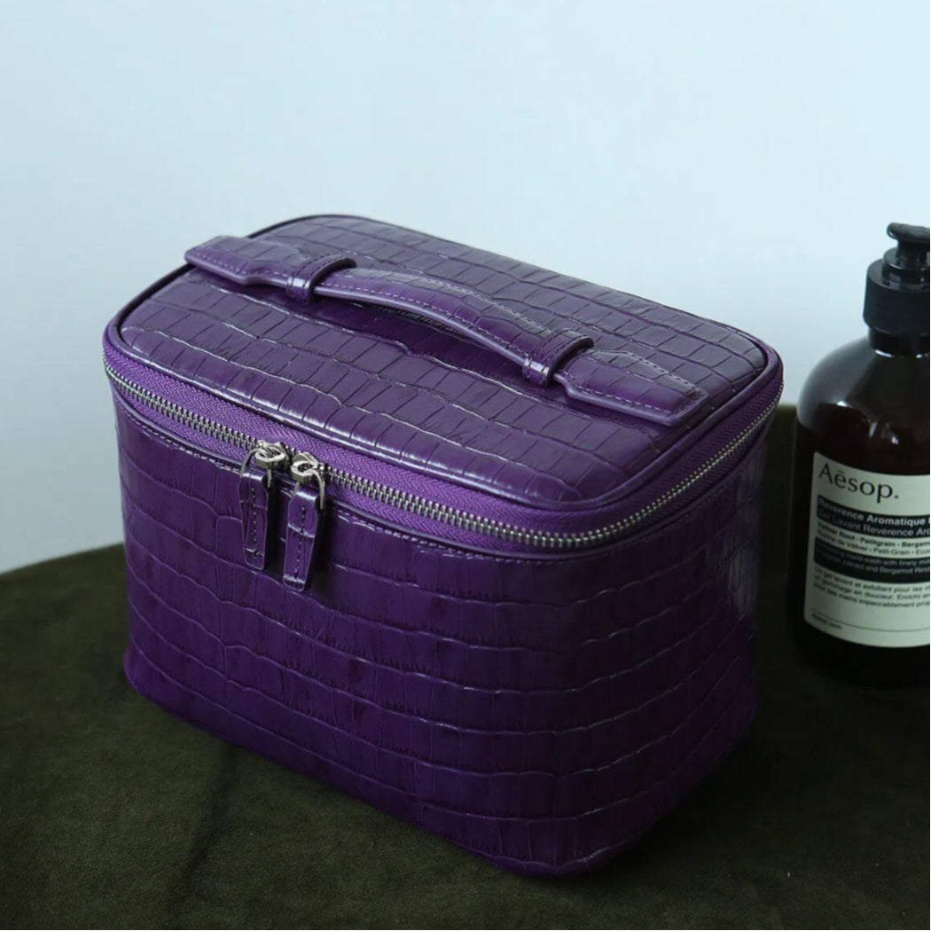 A purple cosmetic bag with a top handle and zipper closure, shown on a dark surface next to an Aesop bottle against a light gray wall.