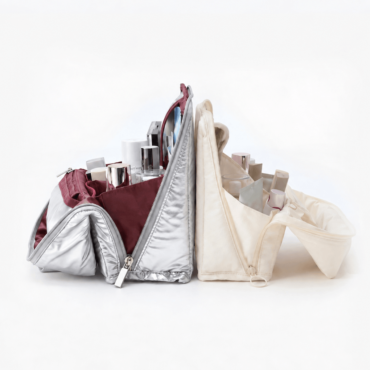 Two zippered cosmetic bags shown open on a clean white background, filled with makeup items. One bag is metallic silver with a burgundy interior, and the other is cream canvas with a gray interior.