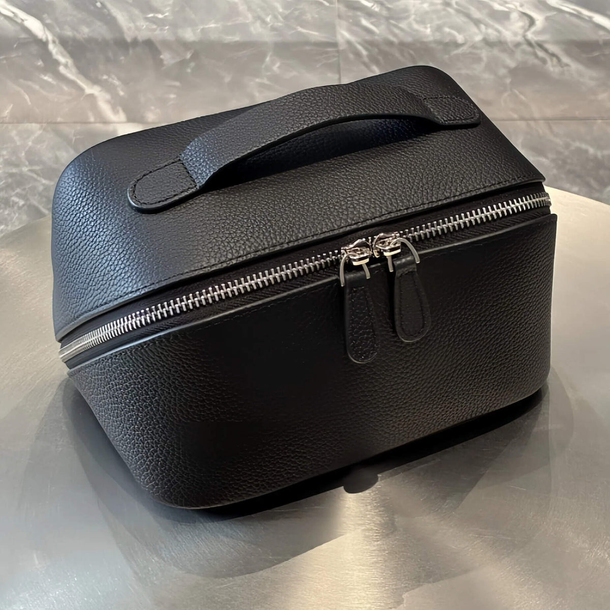 A black pebbled leather cosmetic bag with a top handle and dual zipper closures, resting on a reflective metallic surface.