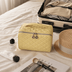 A light yellow quilted cosmetic bag with a silver zipper and dual hanging charms, styled on a white bed alongside a straw hat, an open suitcase, a perfume bottle, a tube of cream, and a makeup brush.