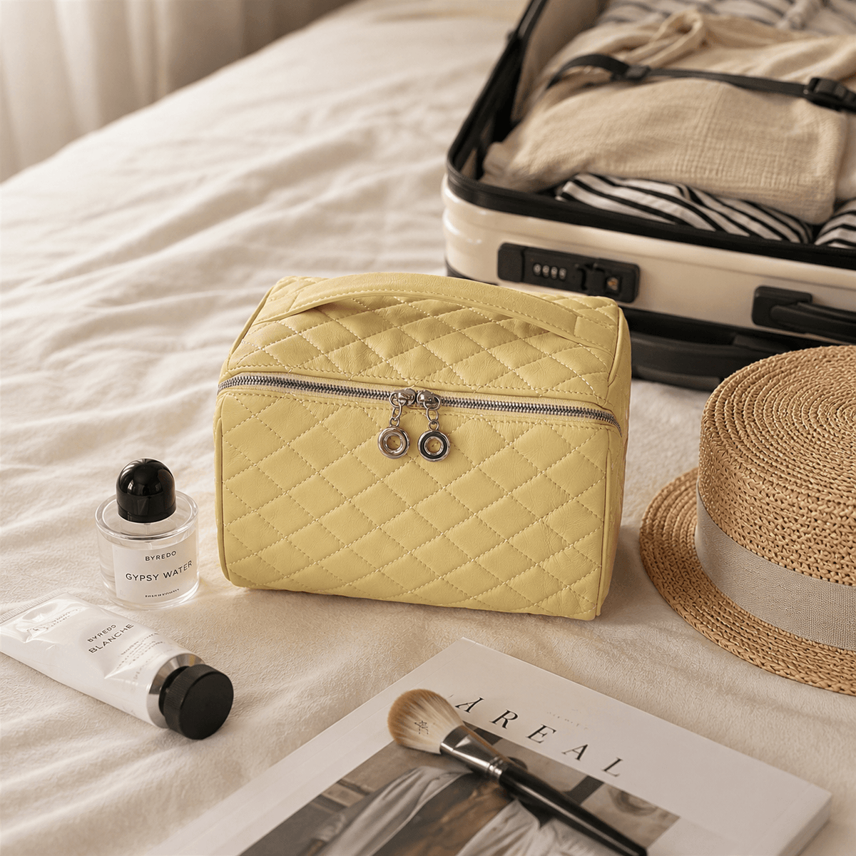 A light yellow quilted cosmetic bag with a silver zipper and dual hanging charms, styled on a white bed alongside a straw hat, an open suitcase, a perfume bottle, a tube of cream, and a makeup brush.