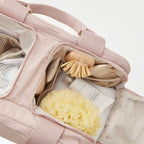 Pink bag with bath products including a scrubber, bottle, and loofah on a light background