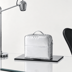 A metallic silver zippered cosmetic pouch with a top handle sits on a black tray on a glass table, with a modern silver desk lamp on the left and a black chair on the right against a white background.