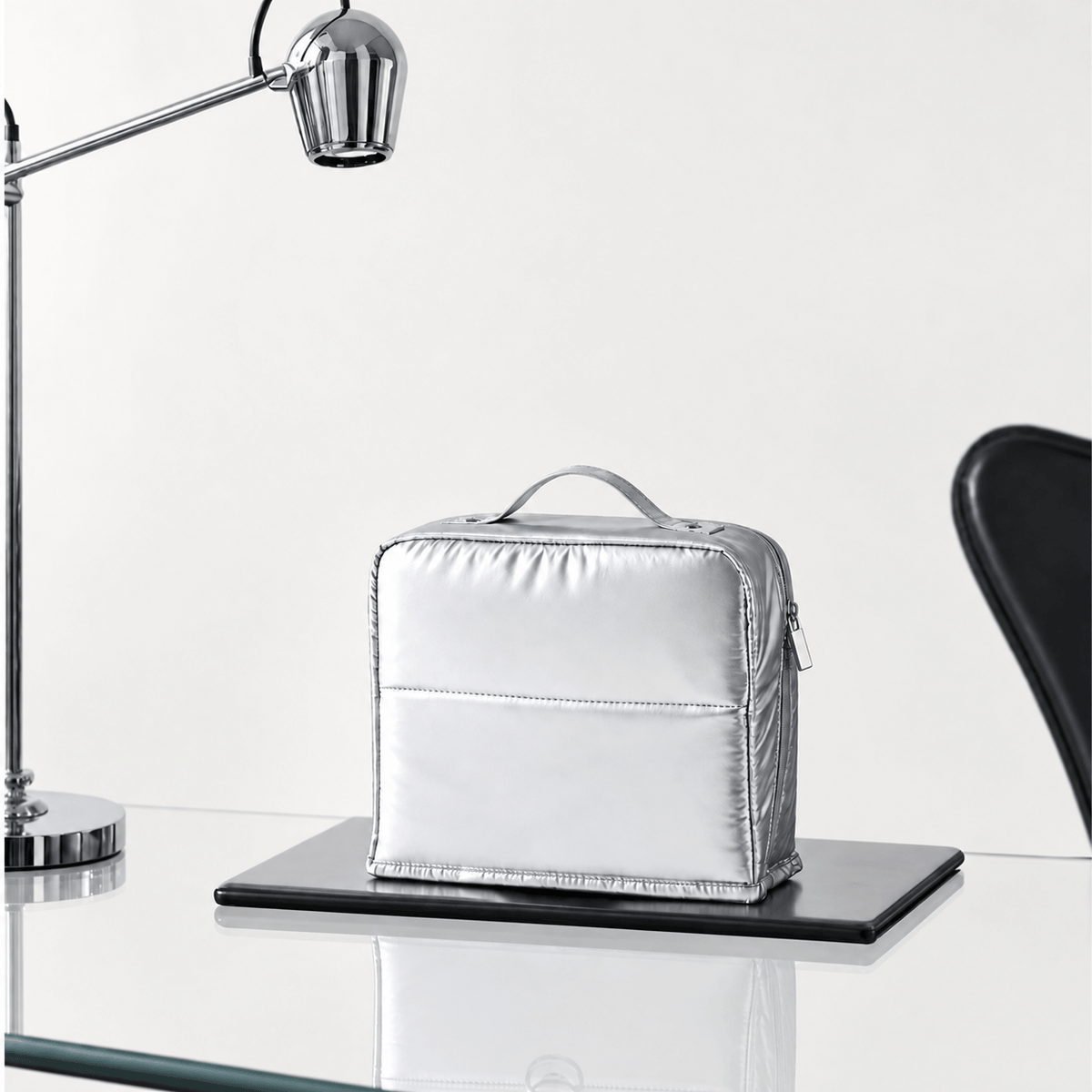 A metallic silver zippered cosmetic pouch with a top handle sits on a black tray on a glass table, with a modern silver desk lamp on the left and a black chair on the right against a white background.