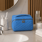 A bright blue quilted cosmetic bag with a top handle and silver zipper sits on a bathroom counter next to a sink, toothbrush holder, and skincare products.