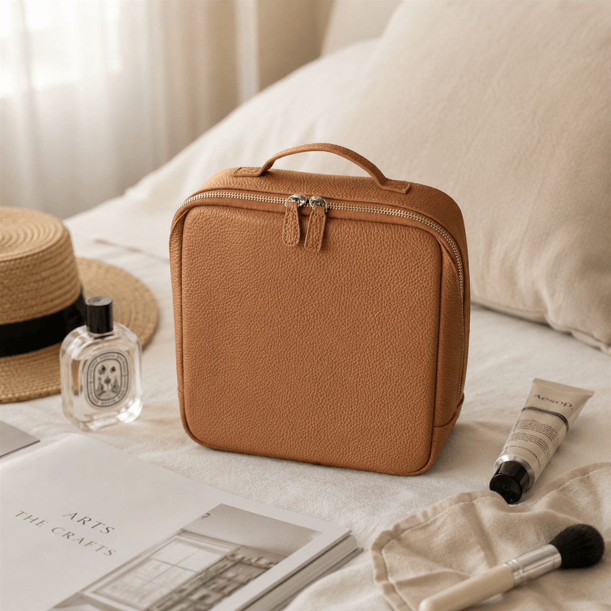 A brown leather cosmetic bag with a top handle and zippered closure, styled on a bed with a straw hat, perfume bottle, skincare tube, and makeup brush nearby.