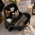 A black zippered makeup bag opened on a marble countertop, filled with cosmetics and skincare items. The bag has a top handle and a clear front pocket.