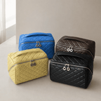 Four quilted zippered cosmetic bags arranged on a light beige surface against a neutral wall. The bags are in blue, mustard yellow, black, and dark brown, each with silver-tone hardware and hanging ring charms.