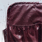 Close-up of a dark burgundy waterproof fabric with water droplets beading on its surface and a ruffled water-repellent seam, shown against a light gray background.