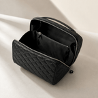 A black quilted cosmetic bag shown open, revealing a black interior with a zippered compartment, photographed on a clean light beige surface with soft shadows.