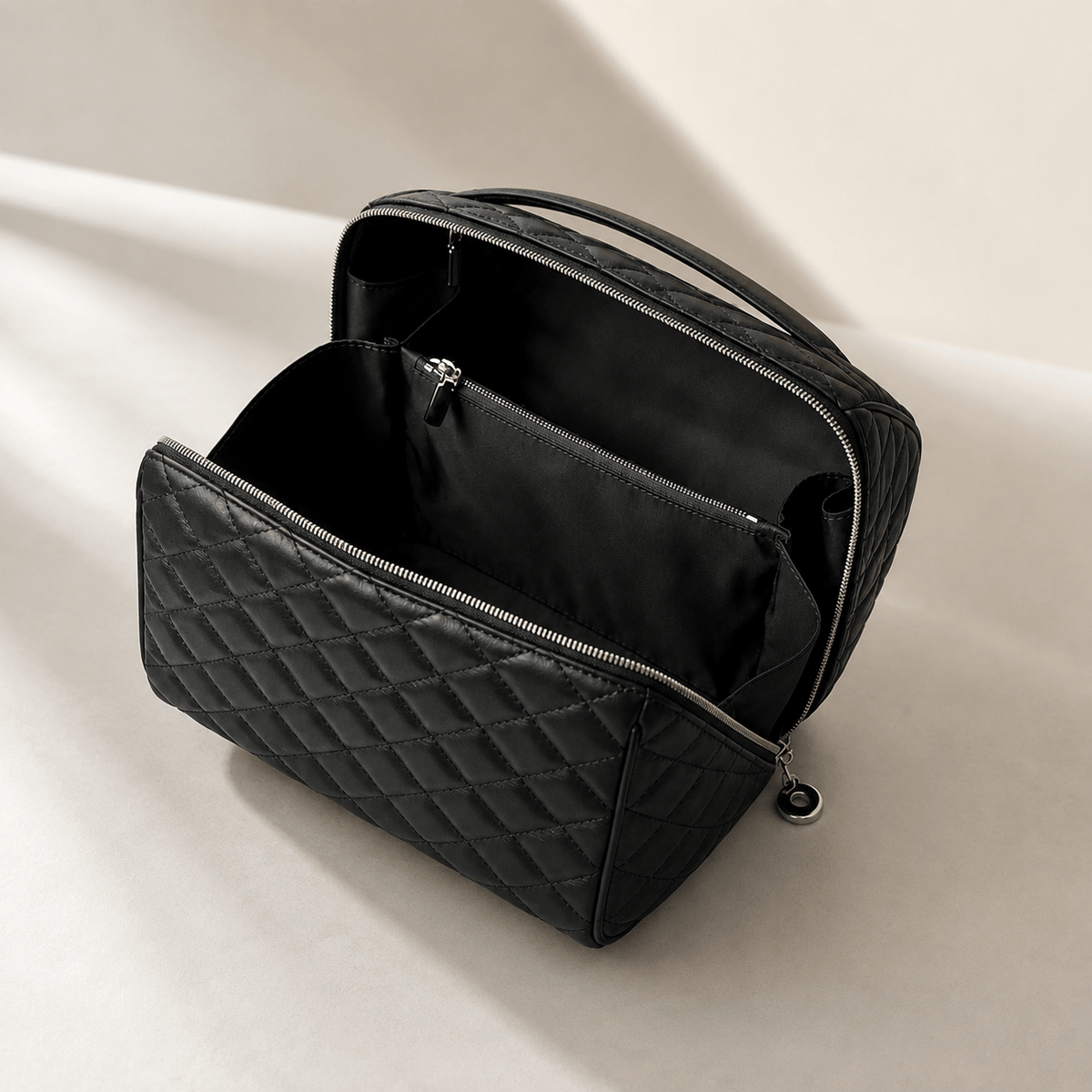A black quilted cosmetic bag shown open, revealing a black interior with a zippered compartment, photographed on a clean light beige surface with soft shadows.