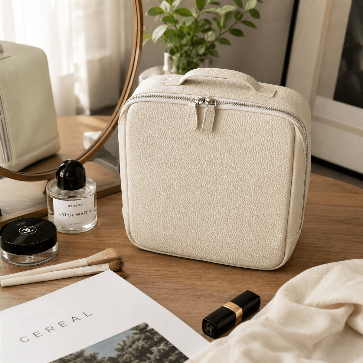 A cream-colored textured leather cosmetic bag with a top handle and zippered closure, styled on a wooden table with cosmetics, a perfume bottle, and a magazine.