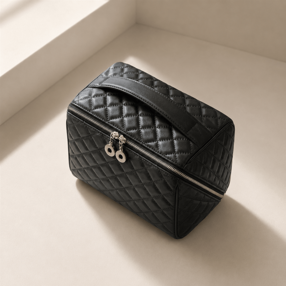 A black quilted cosmetic bag with a top handle, silver zipper, and silver zipper pulls, photographed on a warm beige surface in a minimalist corner setting.