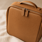 A close-up of a brown leather cosmetic bag with a top handle, dual zipper pulls, and silver-tone zippers, resting on a soft beige fabric surface.