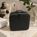 A black pebbled leather cosmetic bag with a top handle and silver zipper, placed on a marble bathroom counter surrounded by skincare and toiletry products.