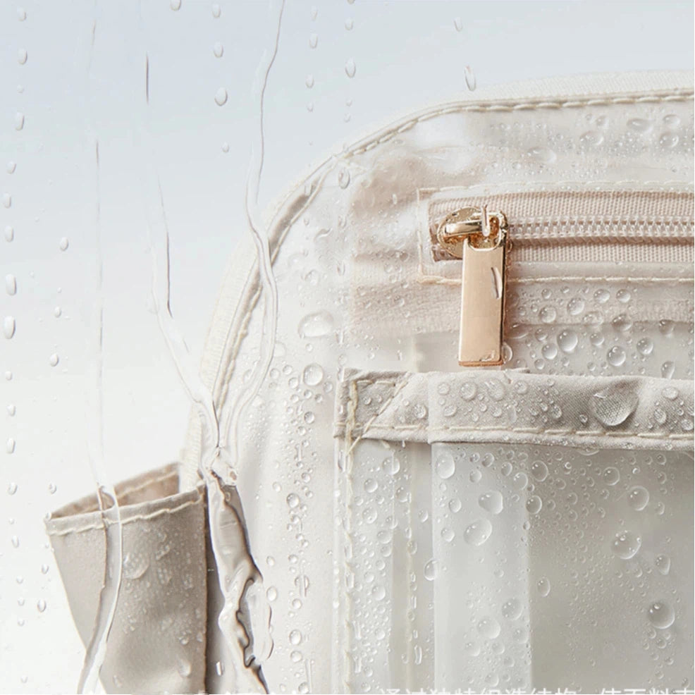 Close-up of a cream cosmetic bag with a transparent water-resistant front pocket covered in water droplets. A metallic gold zipper pull is visible, and the background is white with water streaks and droplets.