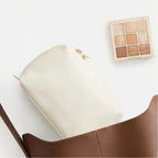 A cream-colored makeup pouch with a gold zipper pull, shown partially inside a brown leather bag next to a nine-shade eyeshadow palette on a clean white background.