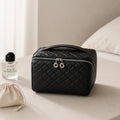 A black quilted cosmetic bag with silver zipper pulls sits on a beige bed next to a perfume bottle and a cream drawstring pouch.