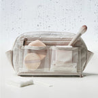 A clear zippered makeup bag with multiple front pockets, shown filled with cosmetic items and a brush, on a white surface with water droplets and a few cotton pads in front.