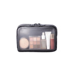 A dark grey zippered cosmetic bag with a clear front window, shown on a plain white background. Inside the bag are several makeup items, including a round compact, a 9-shade eyeshadow palette, a dropper bottle, a white tube, and a red lip product.