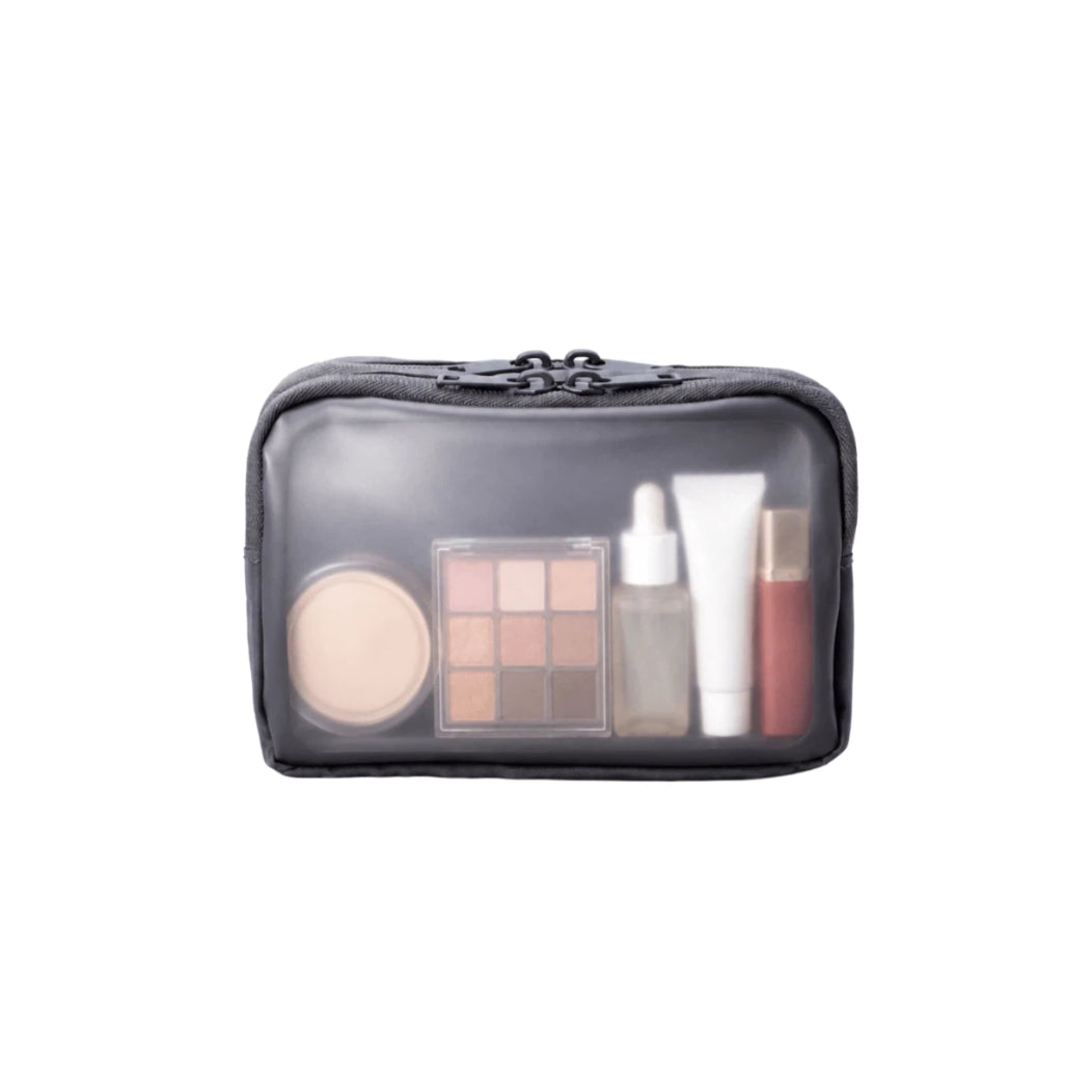 A dark grey zippered cosmetic bag with a clear front window, shown on a plain white background. Inside the bag are several makeup items, including a round compact, a 9-shade eyeshadow palette, a dropper bottle, a white tube, and a red lip product.