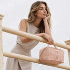 A woman in a beige sleeveless dress leans on a light wood railing, holding a pink quilted cosmetic bag with gold hardware. The background is a clean, bright, overexposed white sky.