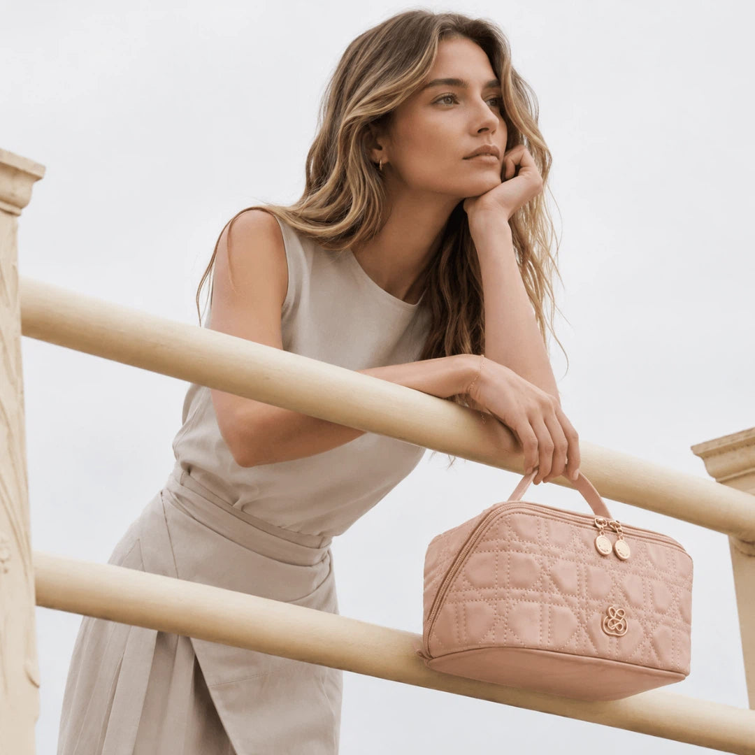 A woman in a beige sleeveless dress leans on a light wood railing, holding a pink quilted cosmetic bag with gold hardware. The background is a clean, bright, overexposed white sky.