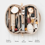 An open beige makeup bag filled with cosmetics and skincare items, arranged in internal compartments. The image includes labeled callouts pointing to different sections: Powders / Touch-ups, Brushes, Lip / Skincare, Makeup, and Samples / Miniatures.