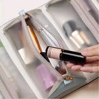 Hand holding a small spray bottle above an opened beige cosmetic bag with makeup items.