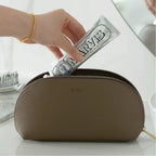 Hand placing a tube of toothpaste into a brown cosmetic bag on a white surface.