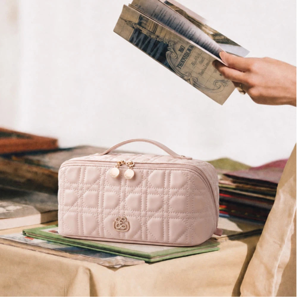 A light pink quilted cosmetic bag with a top handle and gold-tone hardware sits on a tablecloth-covered surface, styled with books and magazines. A hand holding a magazine enters from the upper right.