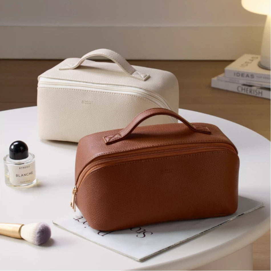 Two cosmetic bags, one beige and one brown, on a table with a brush and small bottle.