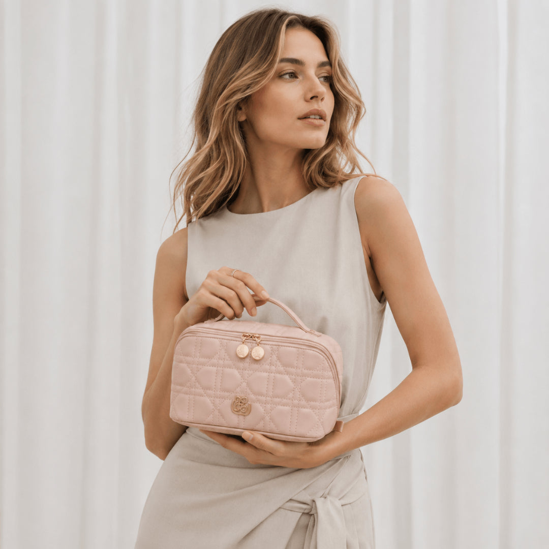 A woman in a light beige sleeveless dress holds a light pink quilted cosmetic case with a top handle and rose gold hardware against a soft white curtain background.