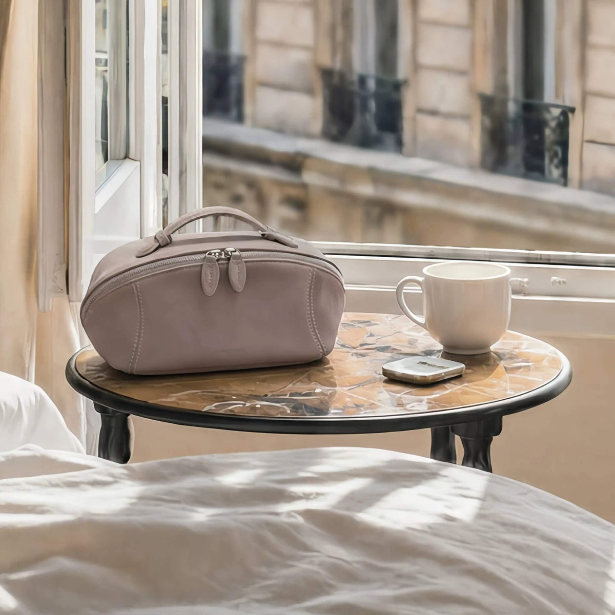 A dusty mauve cosmetic bag with top handles sits on a round marble-topped bedside table next to a white mug and a smartphone, set in a sunlit bedroom with a window and stone building facade in the background.