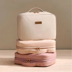 Three stacked cosmetic or travel cases in soft neutral tones: a cream top case with a top handle, a light blush middle case with a side handle, and a dusty pink bottom case, all with gold-tone hardware on a beige studio background.