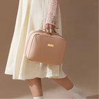 A person in a cream dress and white socks carries a structured blush pink cosmetic bag with a gold-tone logo plaque and top handle against a warm beige studio background.