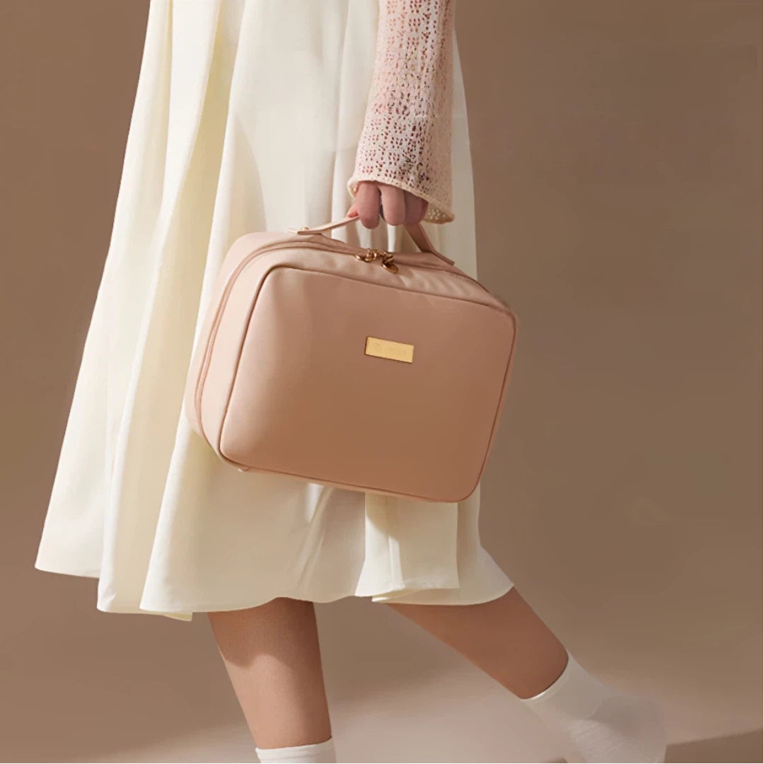 A person in a cream dress and white socks carries a structured blush pink cosmetic bag with a gold-tone logo plaque and top handle against a warm beige studio background.