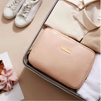 A blush pink zippered travel organizer pouch with a gold logo plaque, neatly packed inside an open suitcase alongside folded cream clothing, white sneakers, and a white towel on a beige background.