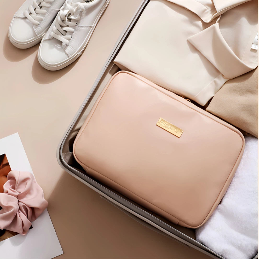 A blush pink zippered travel organizer pouch with a gold logo plaque, neatly packed inside an open suitcase alongside folded cream clothing, white sneakers, and a white towel on a beige background.